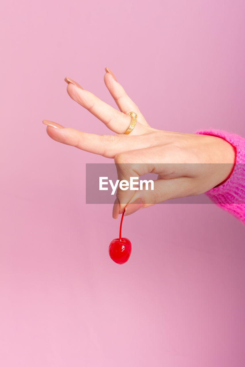 Female hand wearing pink sweater and holding a cherry. light pink background.