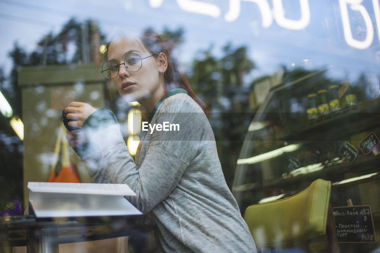 Through glass side view of relaxed female in casual outfit sitting at table with opened book in cozy coffee shop and enjoying aromatic beverage while looking at camera