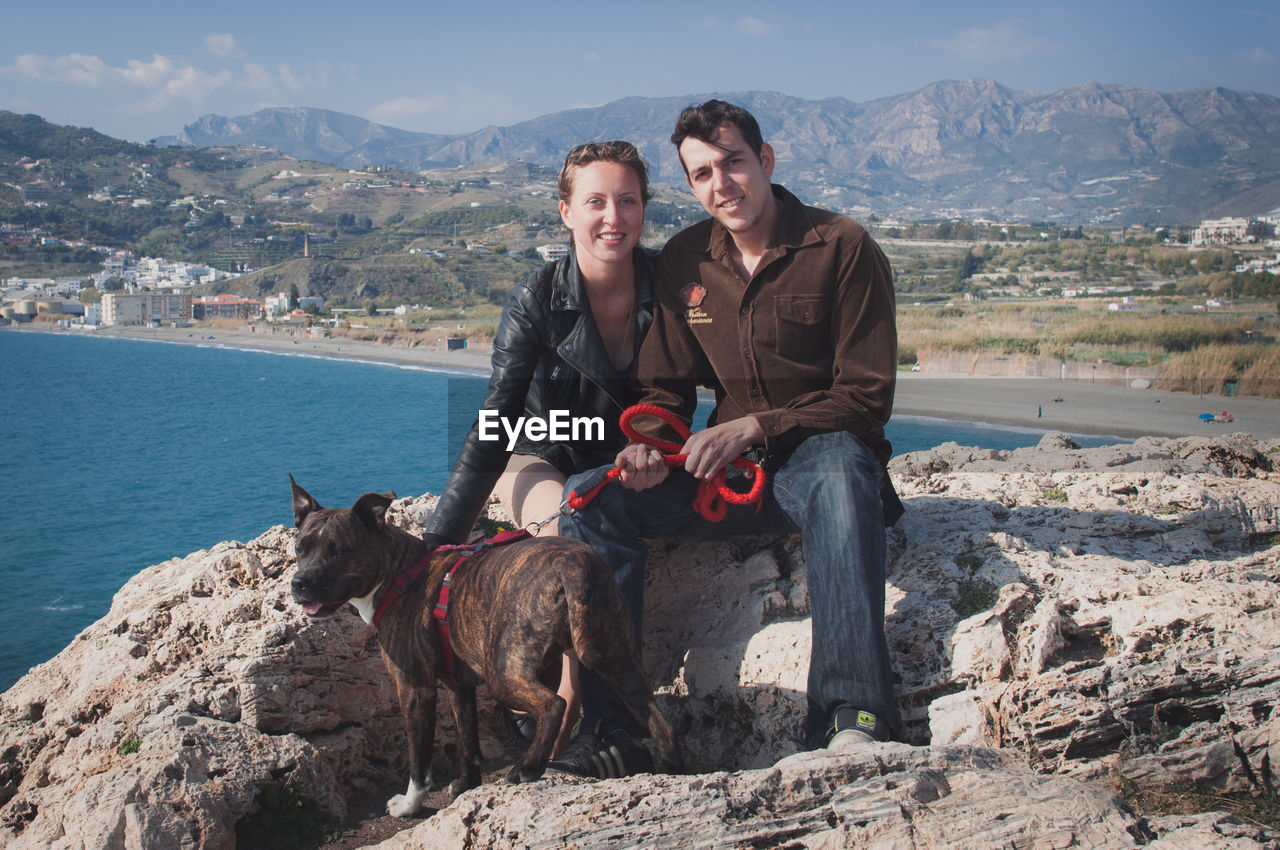 Portrait of couple with dog on rocky mountain against sky