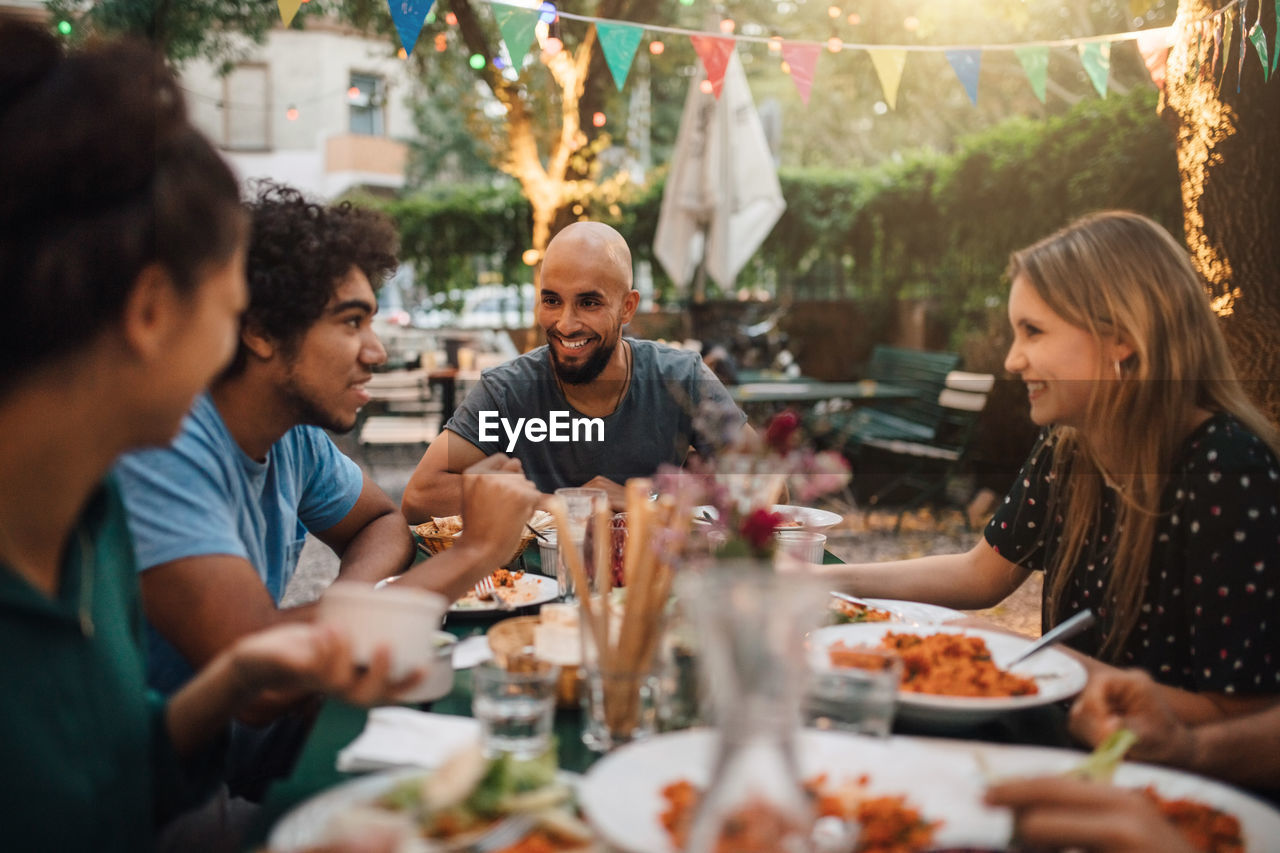 Smiling young man and women listening to male friend talking during dinner party in backyard