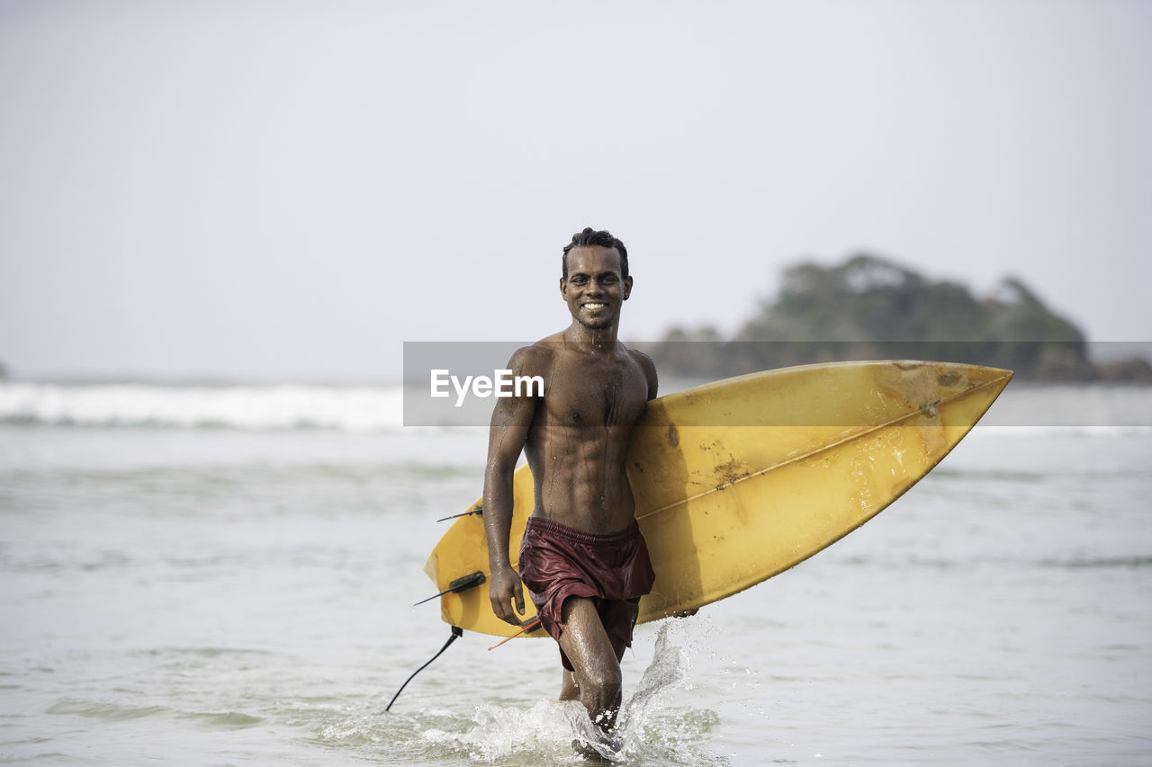 Portrait of shirtless male surfer carrying surfboard in sea