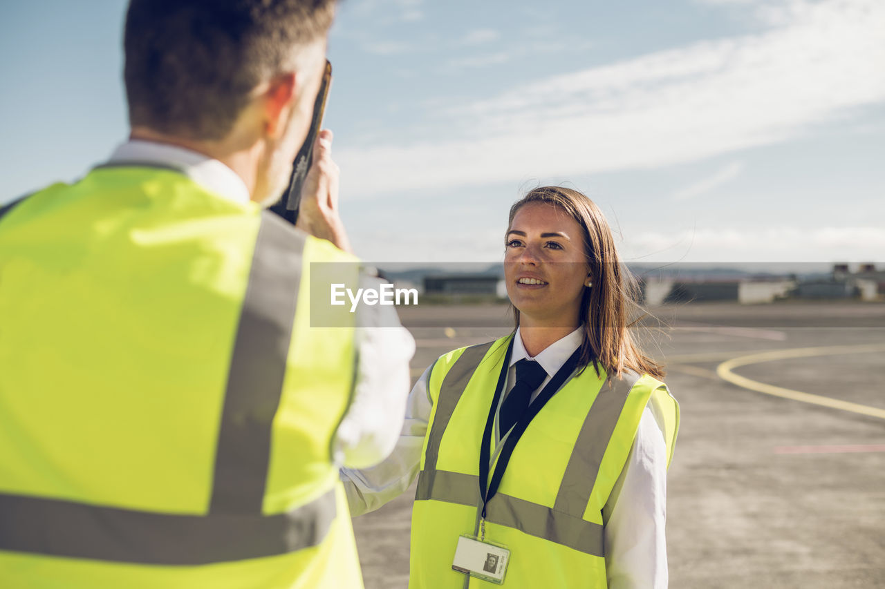 Rear view of male engineer showing airplane parts to female trainee while standing against blue sky on airport runway