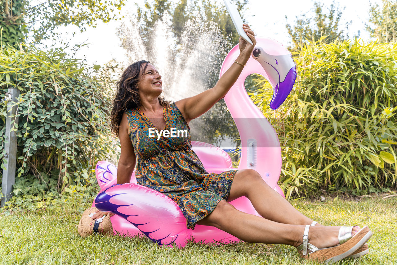 Beautiful smiling middle aged woman taking a selfie sitting on a pink flamingo shaped inflatable toy