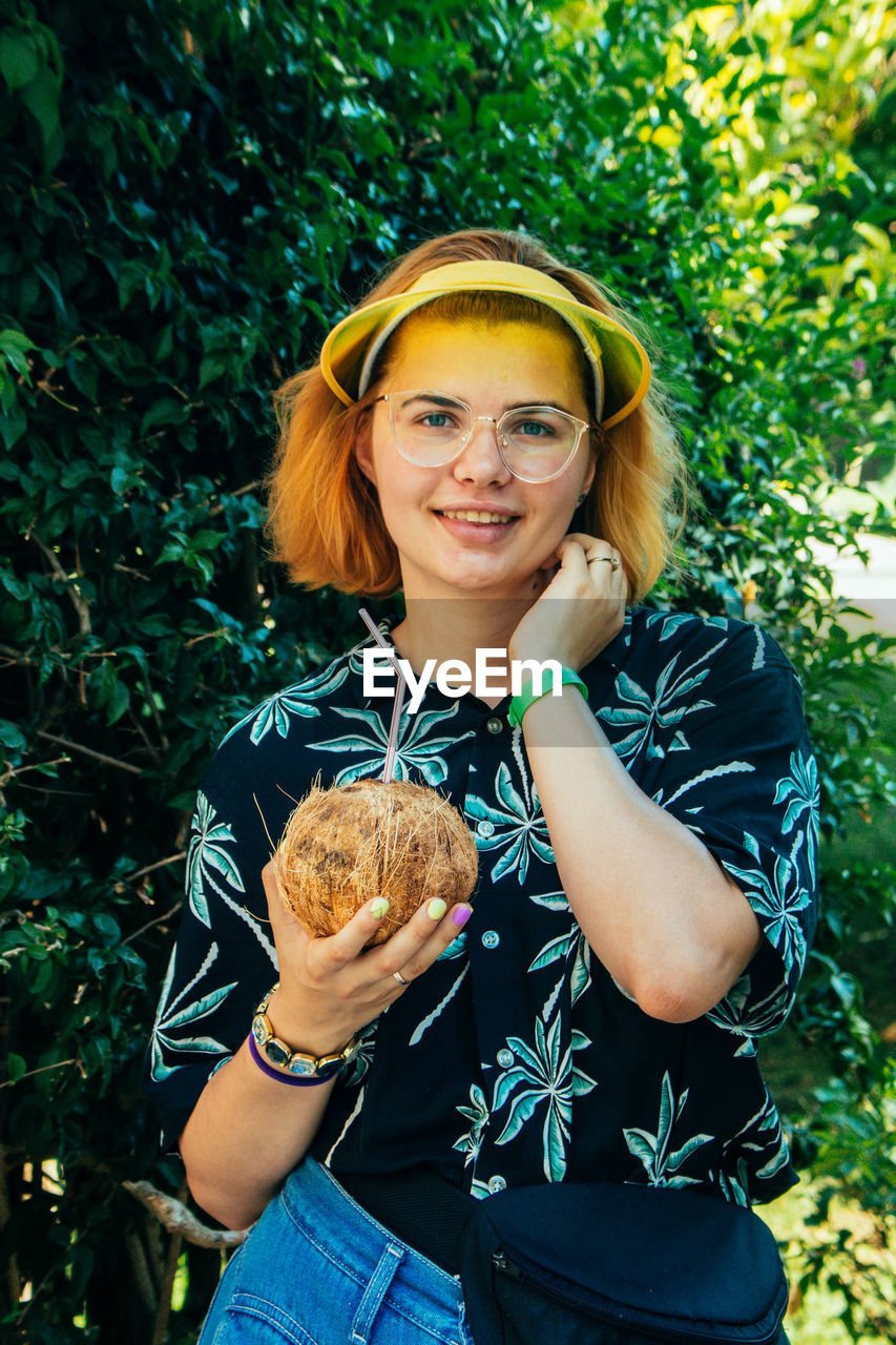Portrait of smiling woman with coconut water standing against trees