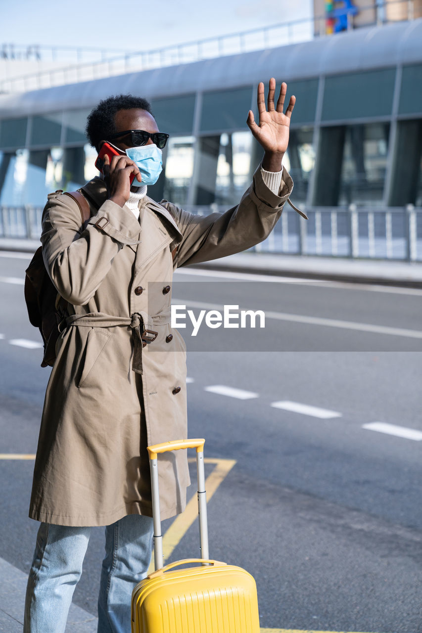 Afro traveler man wear face mask stands in airport terminal, talk on phone, calling taxi. covid-19