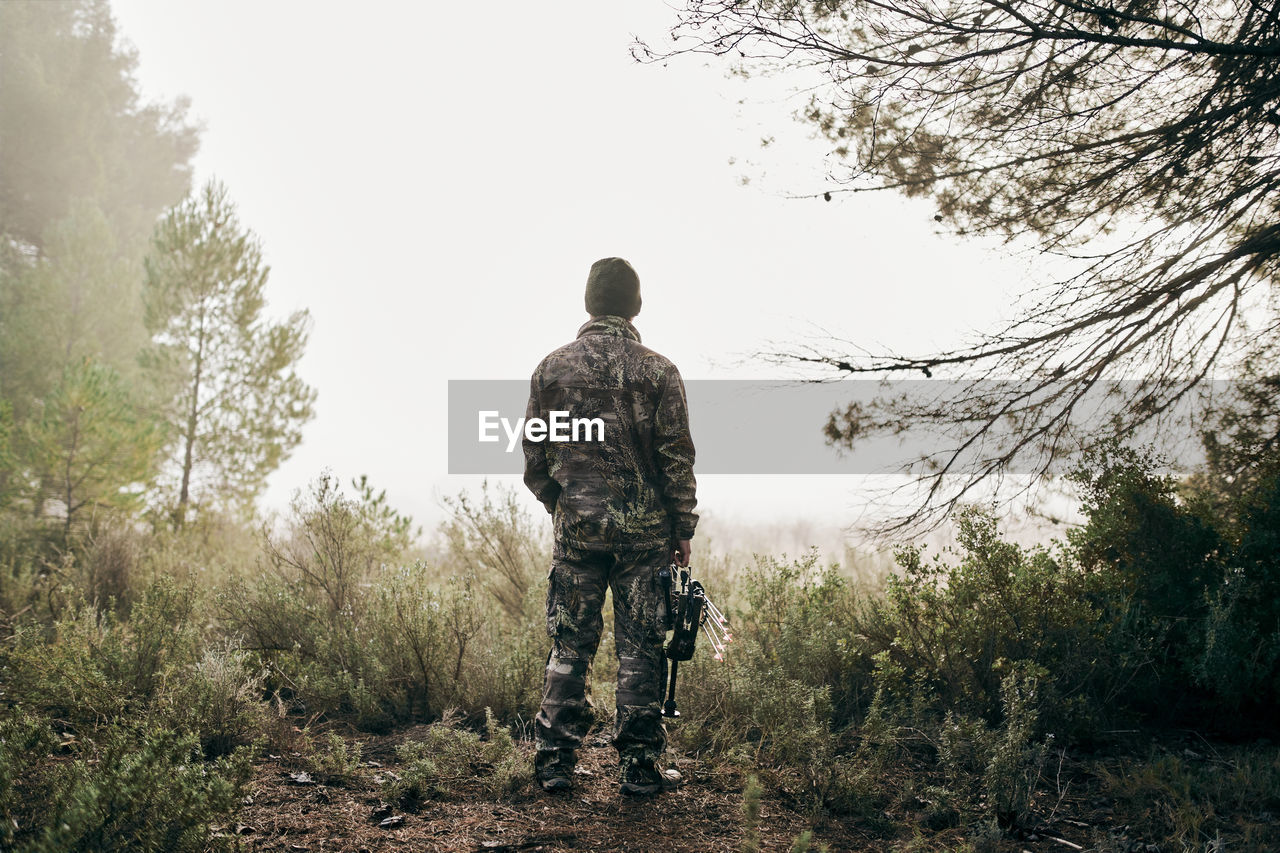 Back view of unrecognizable man in camouflage standing with compound bow in forest and looking away during hunting