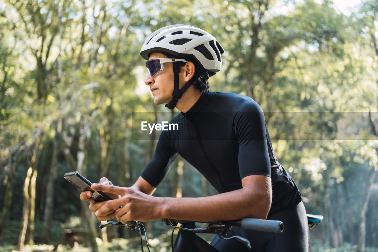 Male biker in helmet and cycling glasses text messaging on cellphone while sitting on bicycle in woods on sunny day