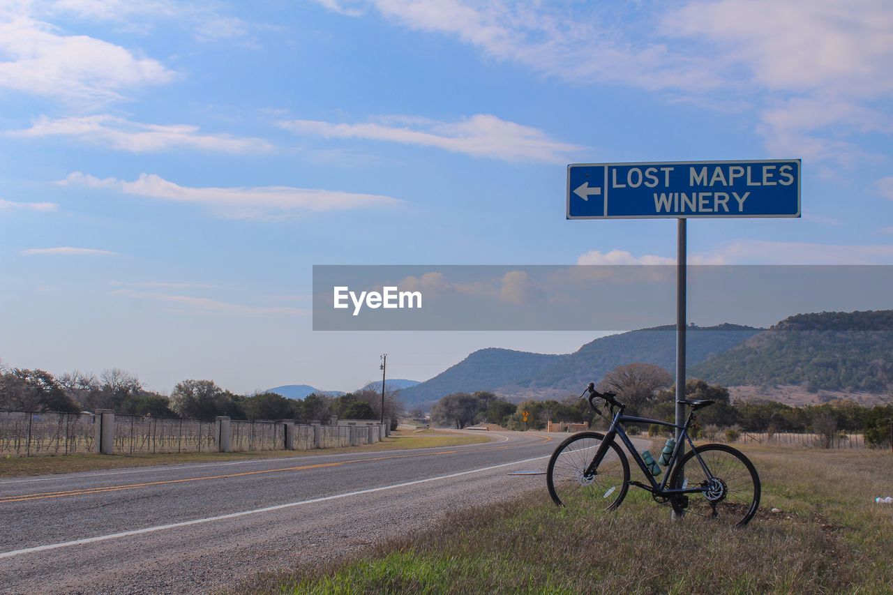 transportation, sign, road, sky, road sign, communication, text, bicycle, western script, nature, cloud, guidance, symbol, no people, landscape, arrow symbol, information sign, vehicle, mode of transportation, travel, day, rural area, highway, environment, lane, directional sign, mountain, outdoors, scenics - nature, land, plant, non-urban scene, infrastructure, rural scene, traffic arrow sign, grass, road trip, tarmac, street, tranquility, tree, cycling, blue