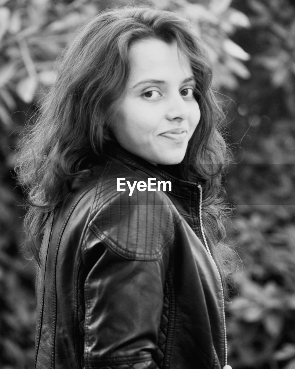 Portrait of smiling young woman wearing leather jacket standing against plants