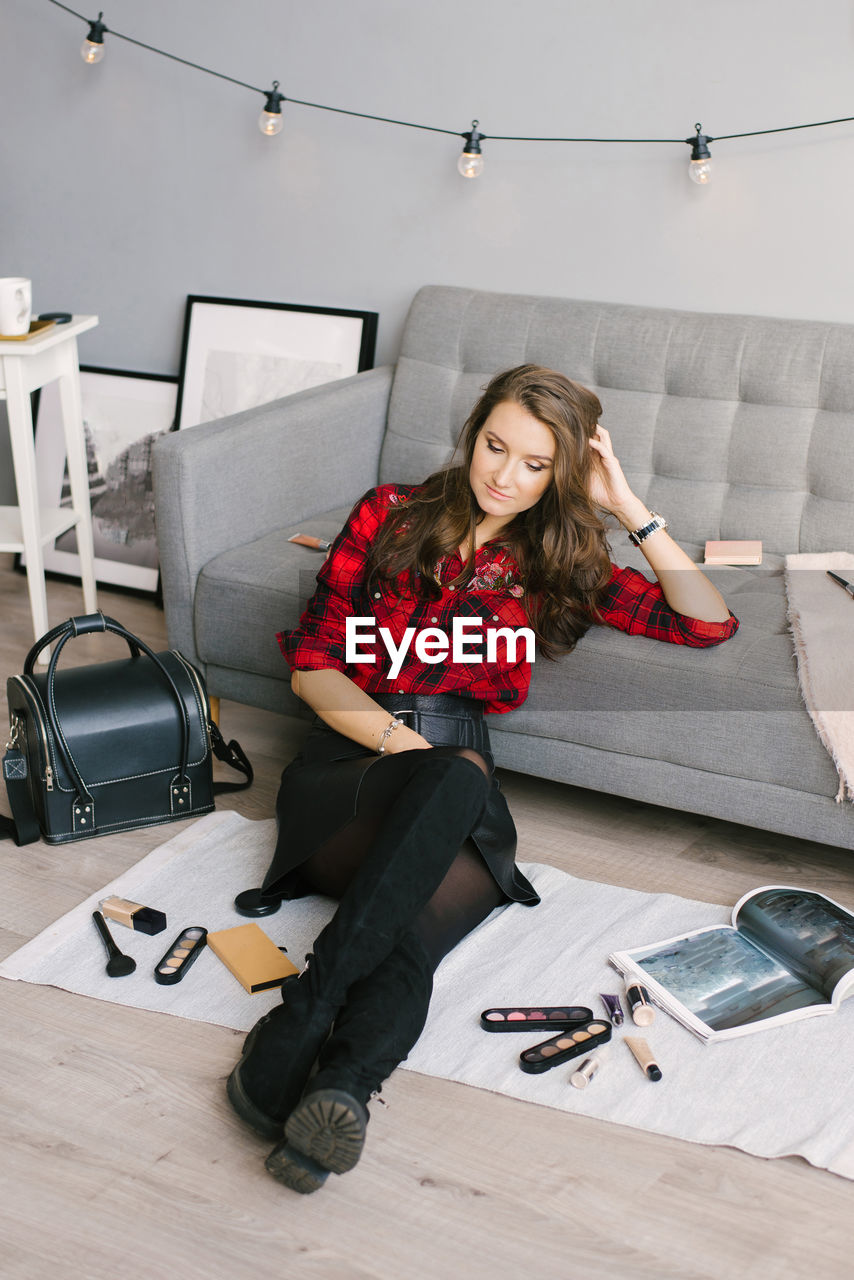 Young female makeup artist is sitting on the sofa floor next to randomly scattered brushes, makeup