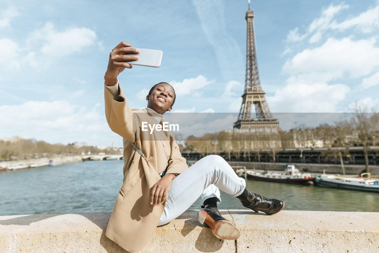 France, paris, woman sitting on bridge over the river seine with the eiffel tower in the background taking a selfie