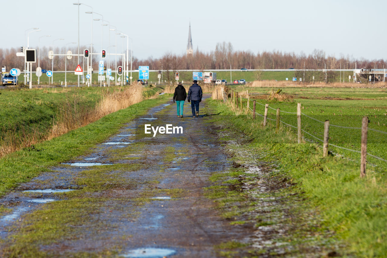 Rear view of people walking on dirt road amidst field
