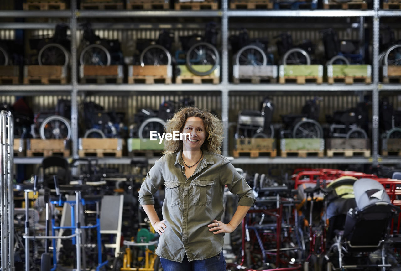 Portrait of smiling mature female worker standing with hands on hip against rack at warehouse