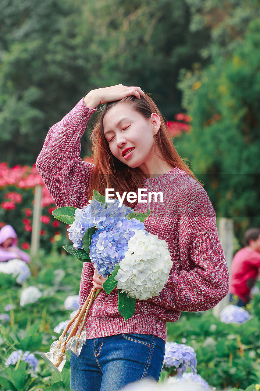 PORTRAIT OF BEAUTIFUL YOUNG WOMAN HOLDING FLOWER IN STANDING AGAINST PLANTS