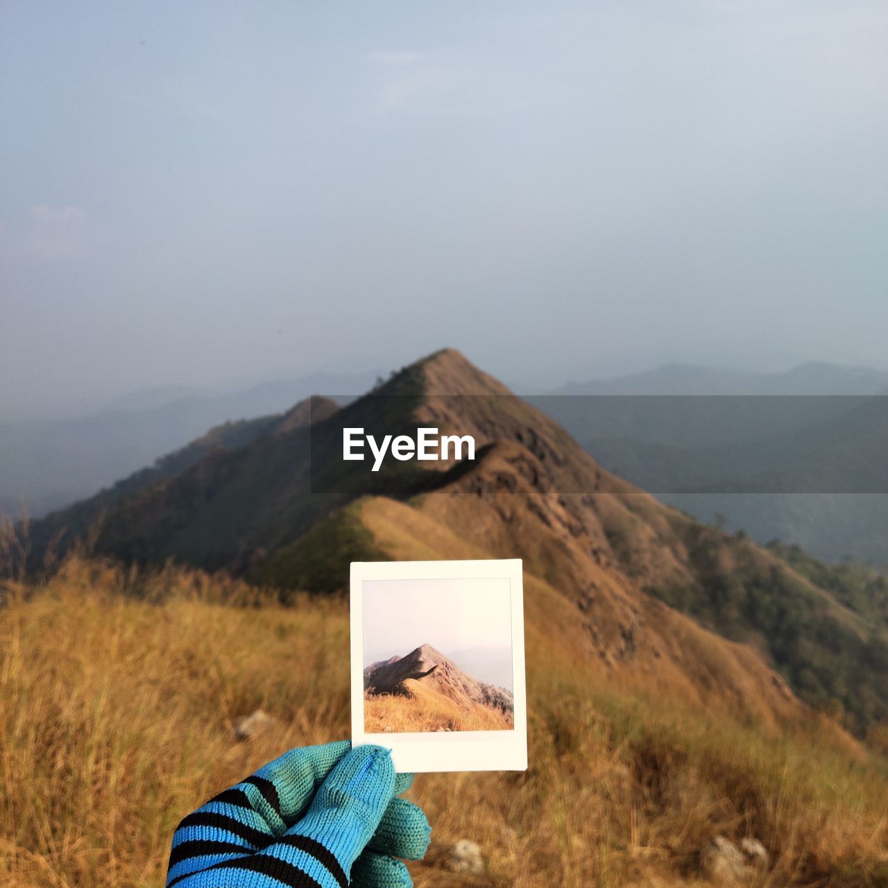 Cropped hand of man holding photograph against mountain and sky