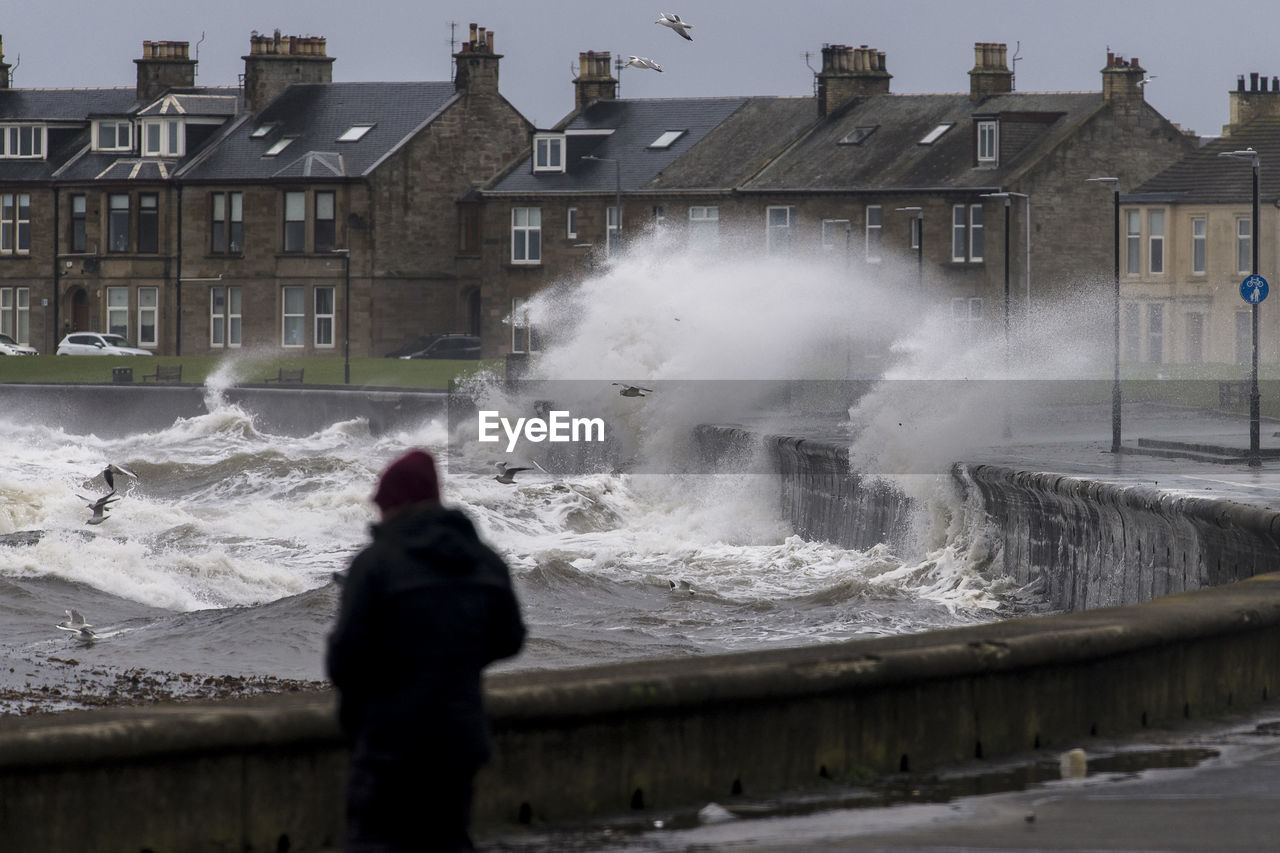 REAR VIEW OF PERSON STANDING IN SEA AGAINST BUILDINGS IN CITY