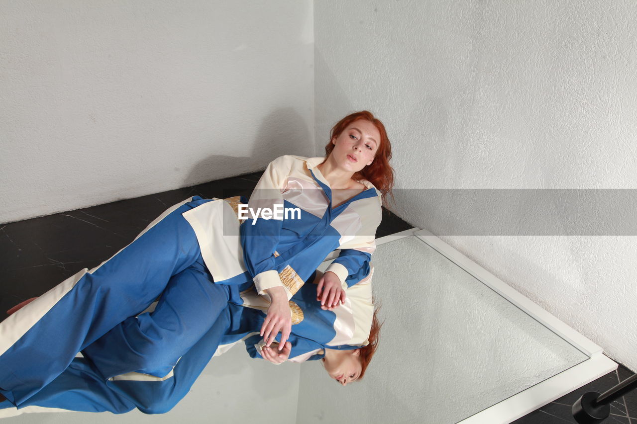 Fashion portrait of ginger woman in fashionable stylish summer suit sitting on mirror, studio