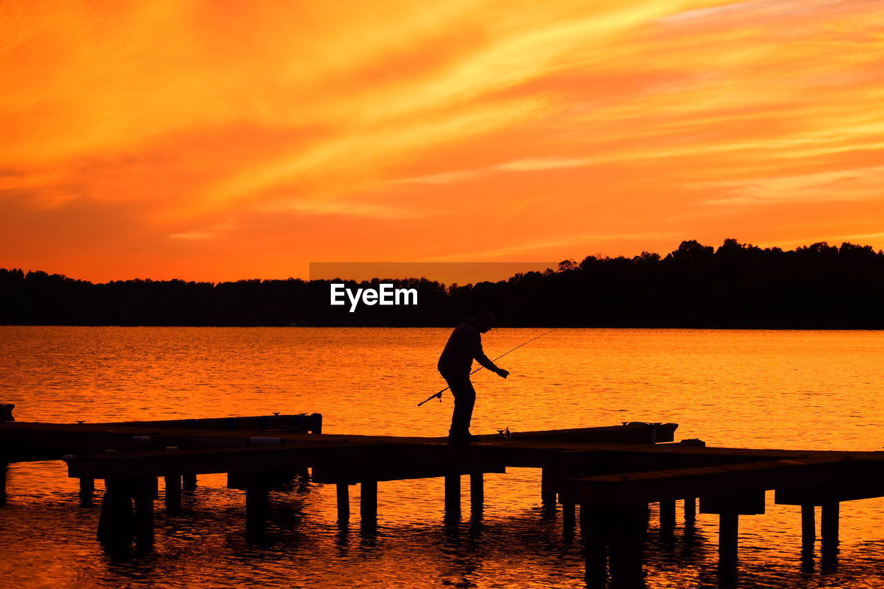 Silhouette man standing by lake against orange sky