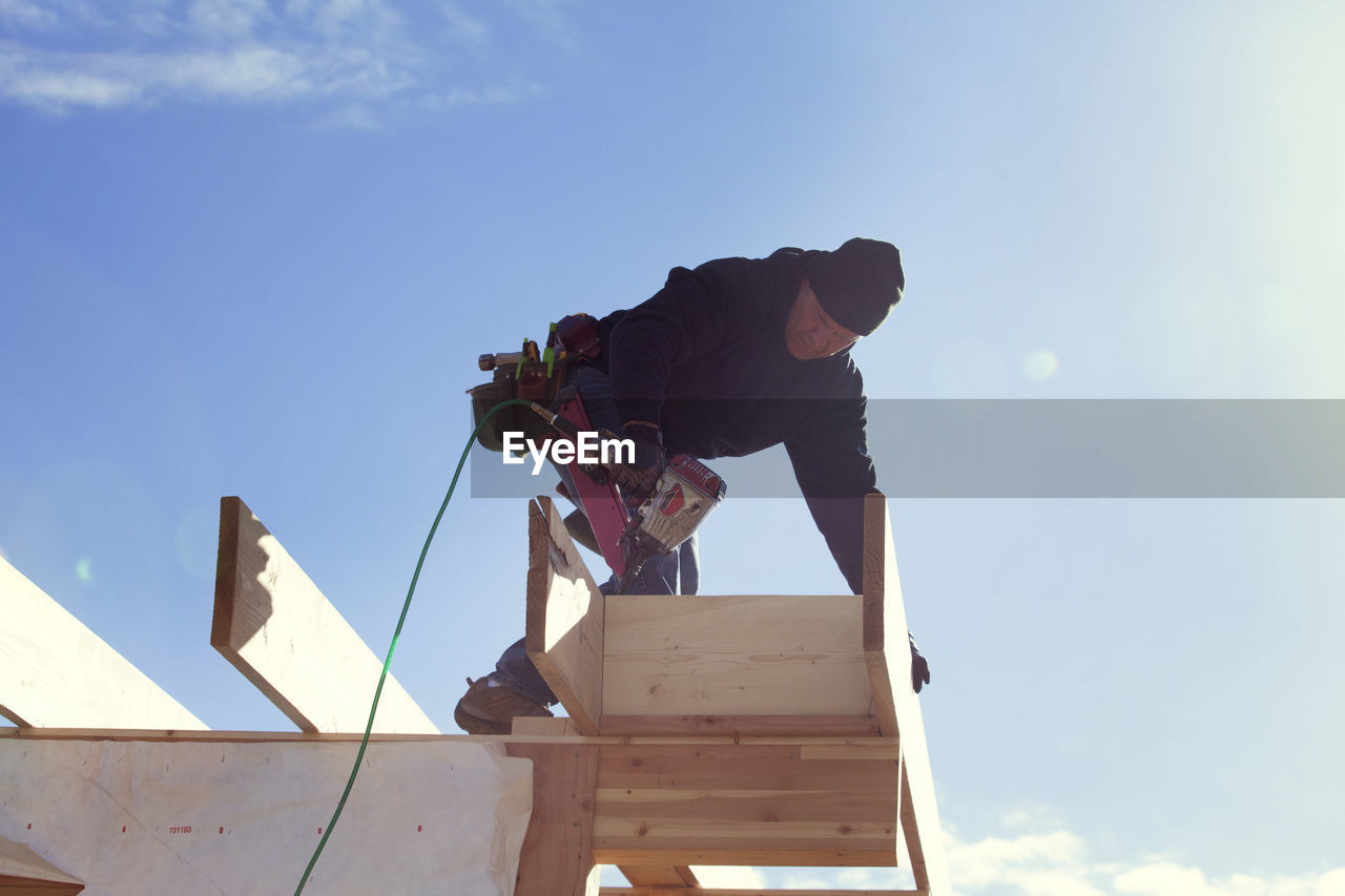 Low angle view of worker with drill standing on roof beam against sky