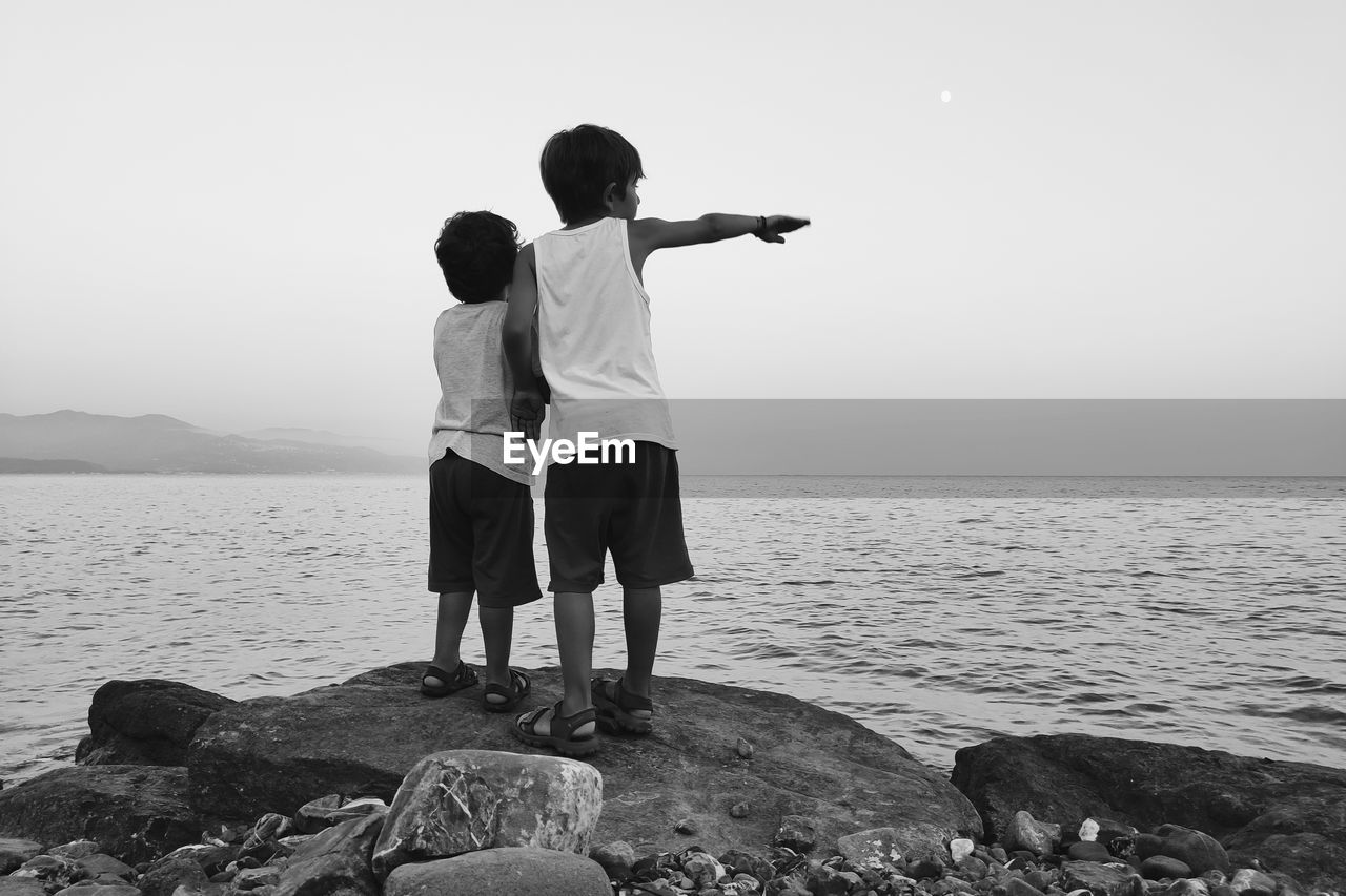 REAR VIEW OF MEN STANDING ON ROCK AT SHORE AGAINST SKY