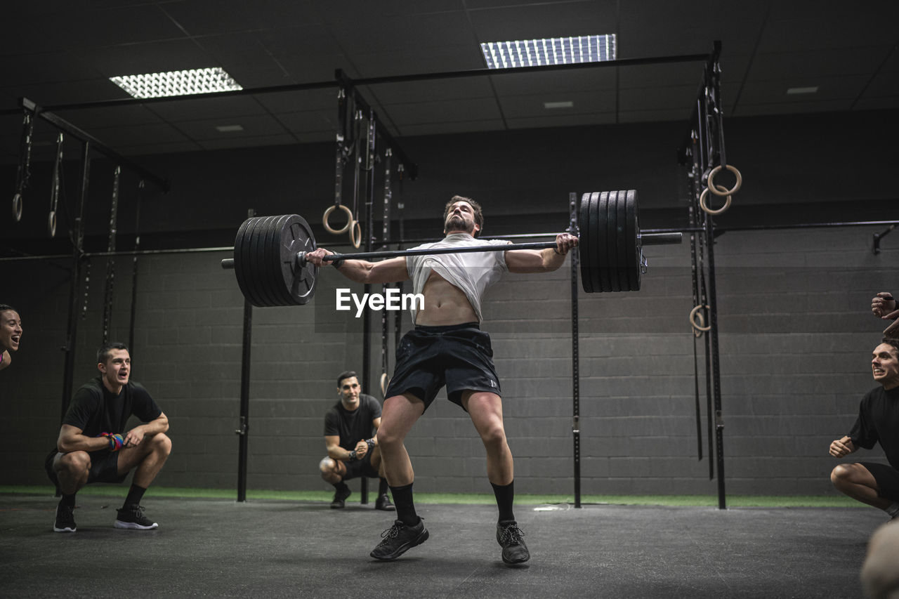 People cheering man lifting barbell while standing in gym