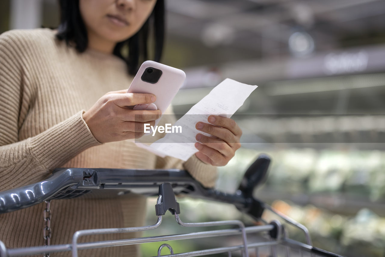 Young woman examining list while using smart phone at supermarket