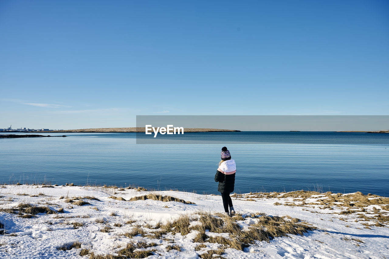 Unrecognizable lady standing on snowy seashore against cloudless blue sky