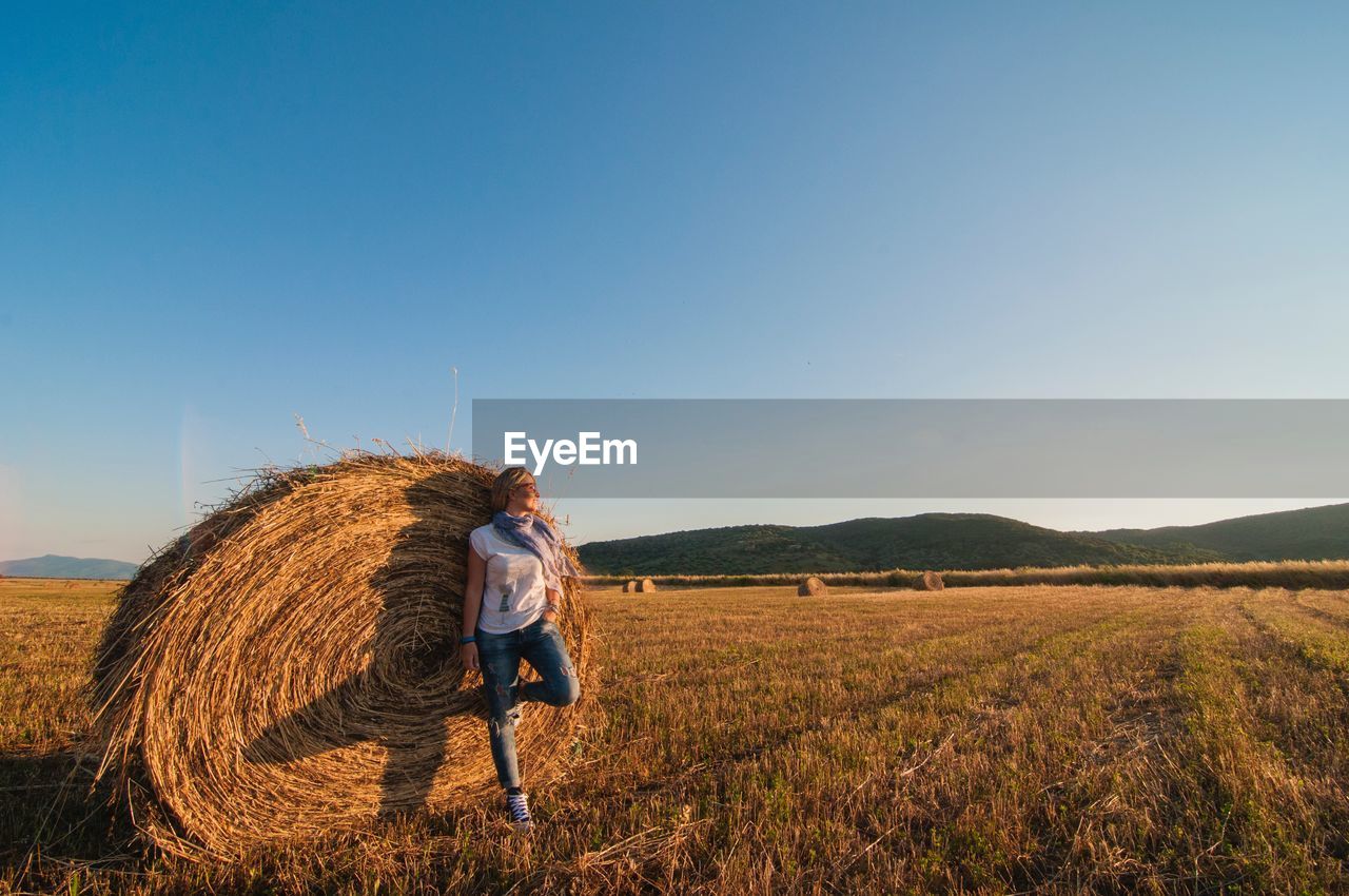 FULL LENGTH OF MAN WEARING HAY BALES ON FIELD AGAINST CLEAR SKY