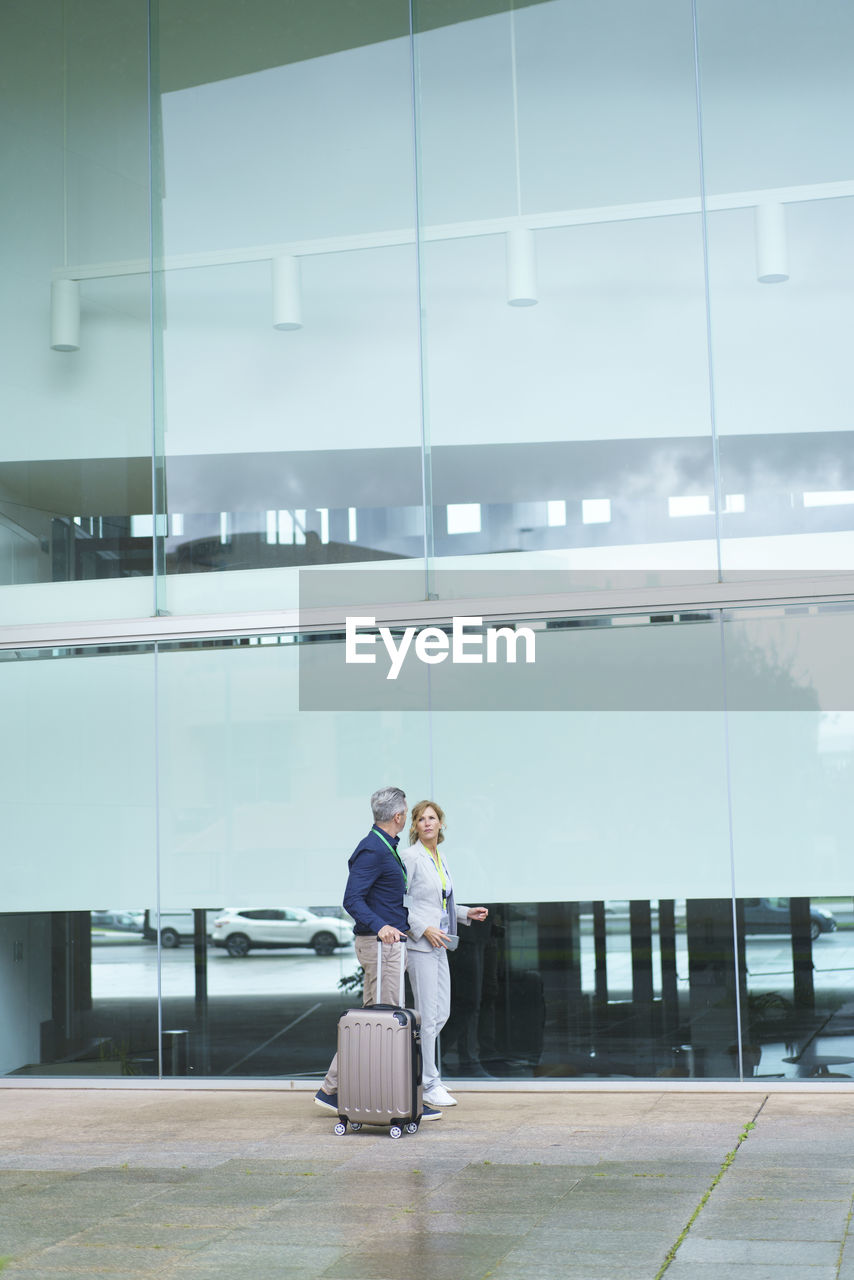 Businessman with suitcase walking by businesswoman in front of glass wall