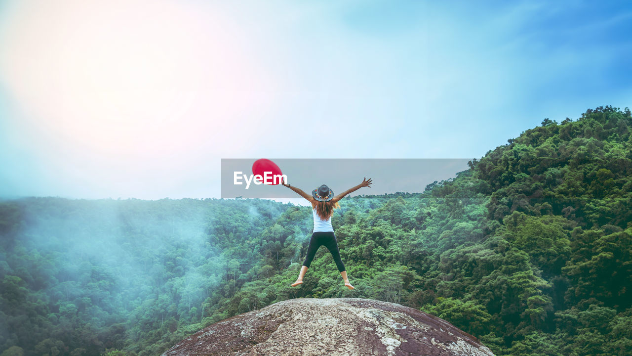 one person, nature, mountain, sky, adult, leisure activity, full length, arm, adventure, rock, environment, plant, arms raised, day, tree, young adult, beauty in nature, land, limb, sports, emotion, outdoors, standing, balloon, women, scenics - nature, lifestyles, copy space, landscape, vacation, vitality, activity, forest, tranquility, trip, extreme sports, human limb, holiday, positive emotion, happiness, sunlight, casual clothing, carefree, person, travel, red, arms outstretched, motion, relaxation, blue, enjoyment, mid-air