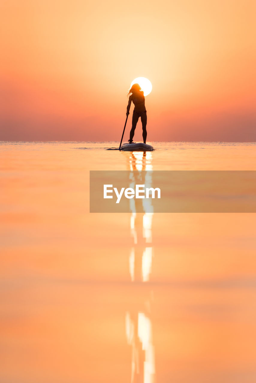 Back view of silhouette of unrecognizable female surfer standing on paddleboard and rowing against spectacular sun in sunset sky