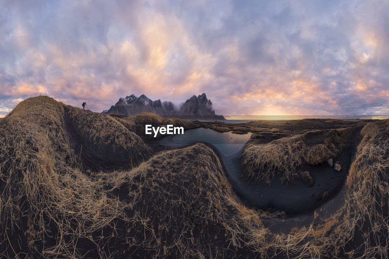 Distant traveler standing on top of mountain range against cloudy sky during sunset near black sand beach in iceland