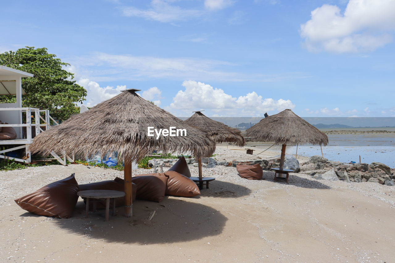 thatched roof, land, sky, beach, vacation, nature, water, roof, sand, sea, chair, cloud, trip, holiday, hut, architecture, beauty in nature, tranquility, day, relaxation, sunlight, thatching, parasol, lounge chair, summer, scenics - nature, travel destinations, beach umbrella, tranquil scene, built structure, umbrella, outdoors, shore, seat, coast, sunshade, building exterior, idyllic, no people, ocean, tropical climate, shade, travel, building, body of water, beach hut, tree, furniture, plant, house, tourism, protection