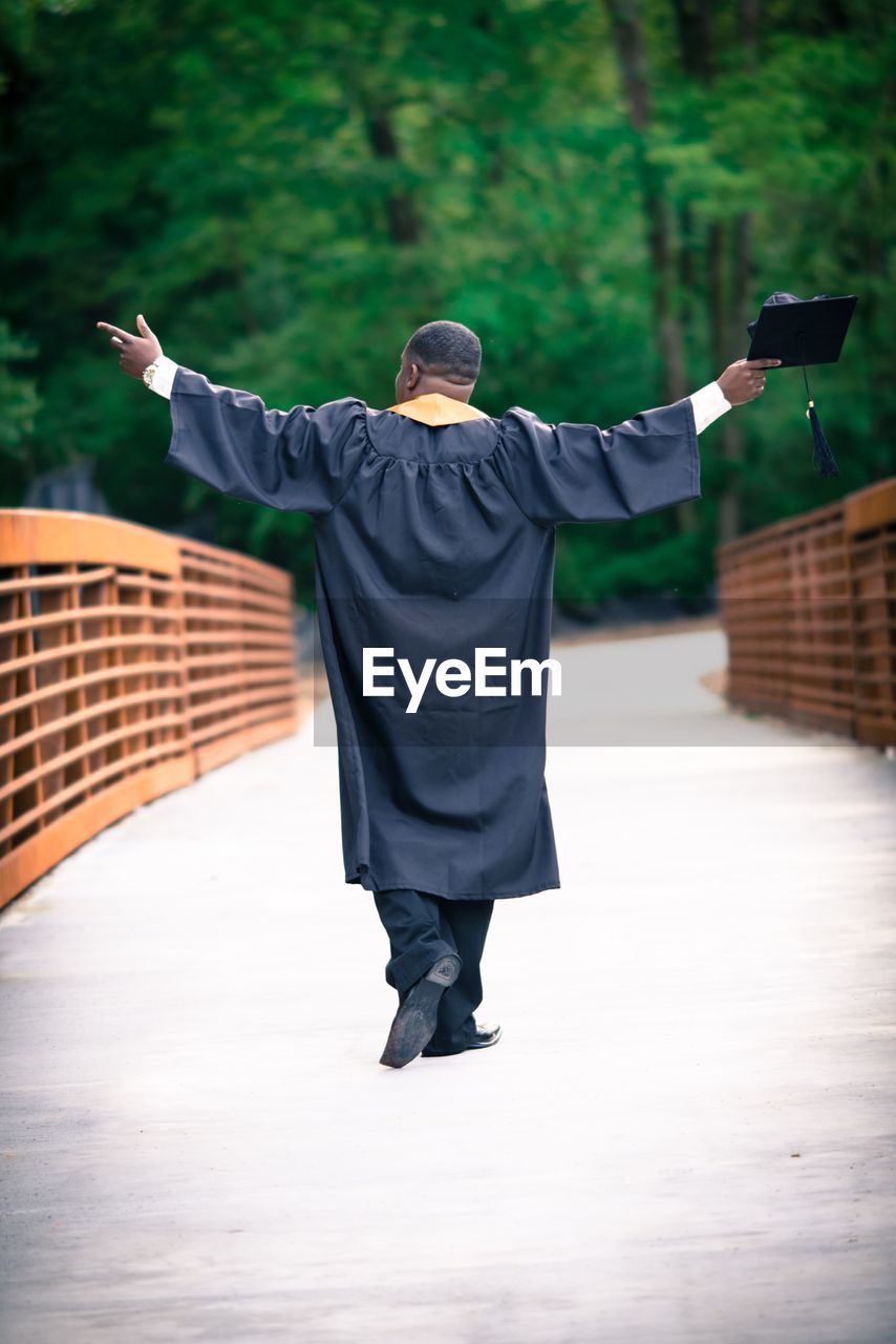 Rear view of man with arms outstretched wearing graduation gown walking on footbridge