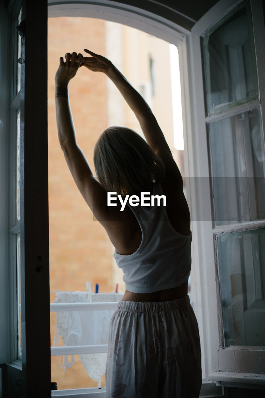 Back view of anonymous female in pajamas standing near window and stretching after awakening at home while enjoying morning