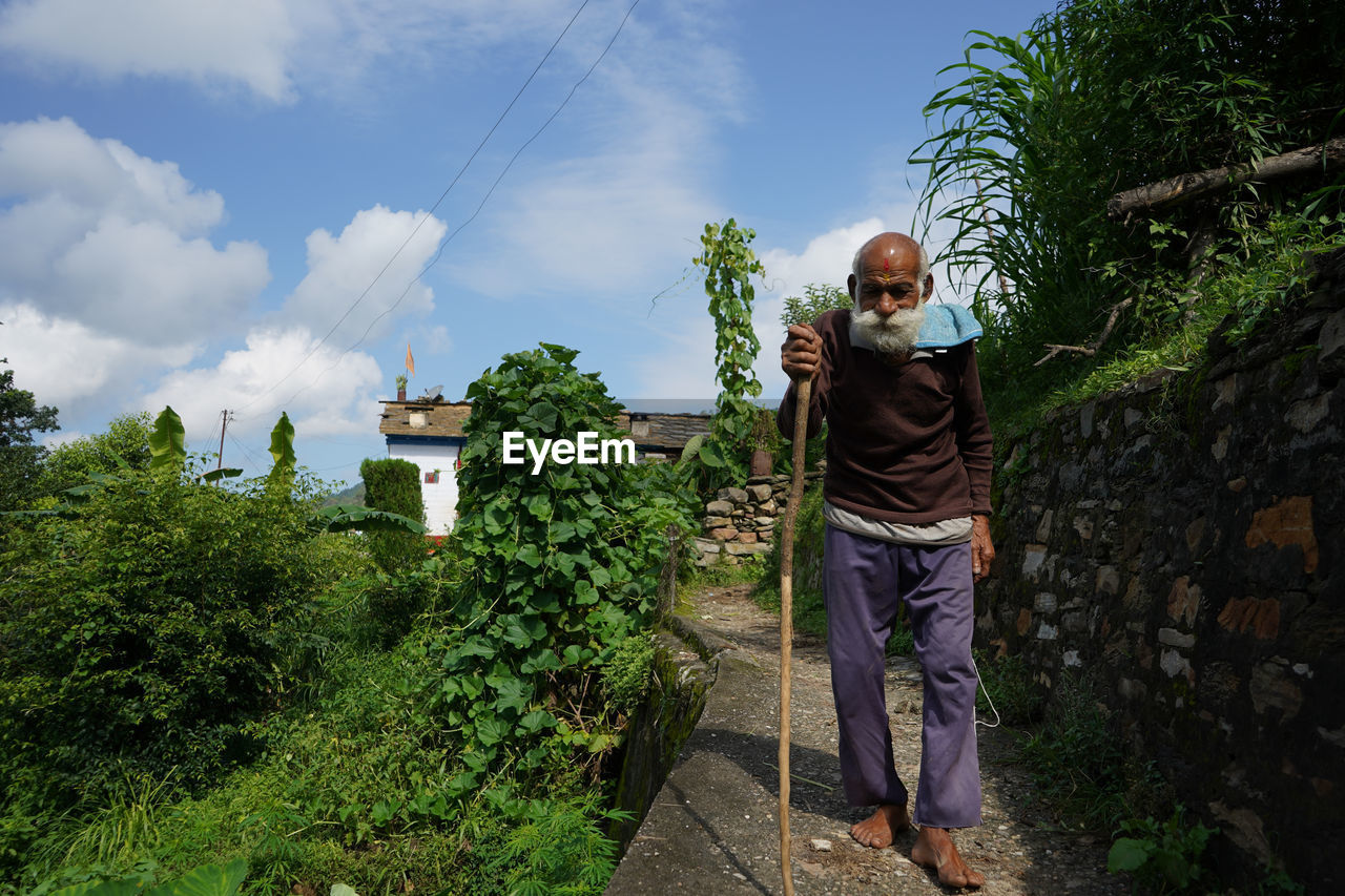 An old man with long white beard walking down a narrow pathway with the help of a stick.