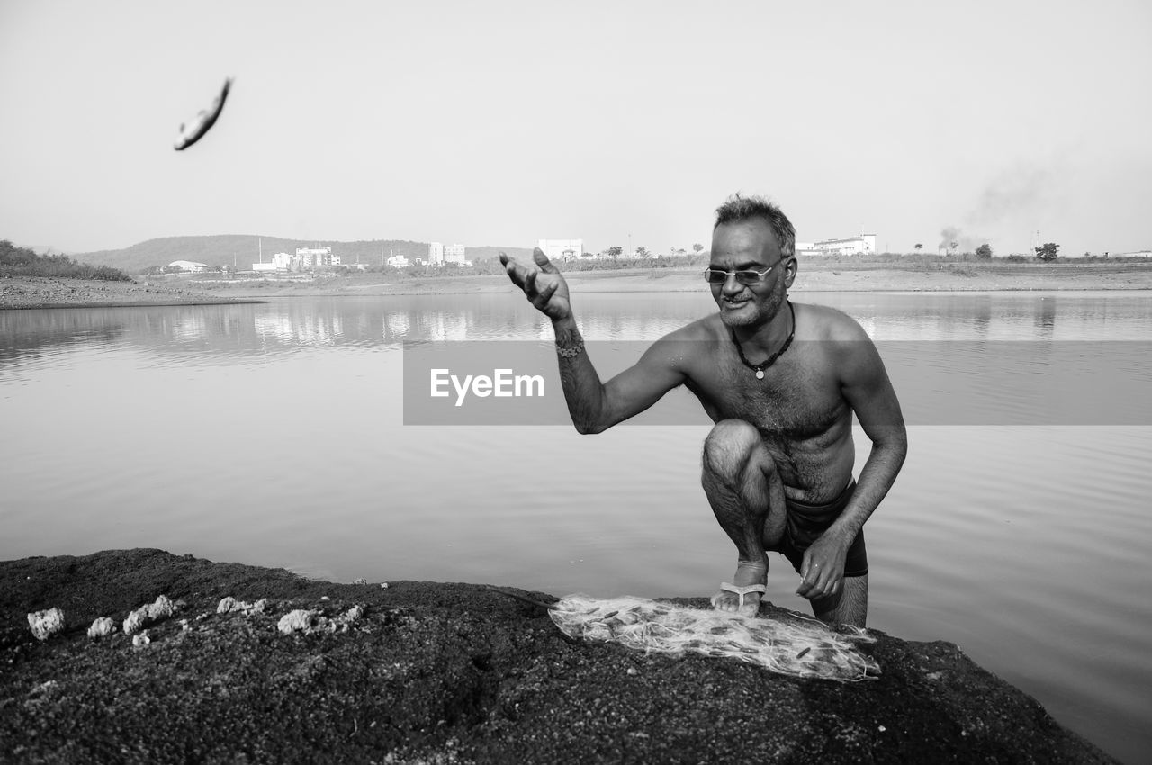 Shirtless fisherman throwing fish from net while kneeling on lakeshore