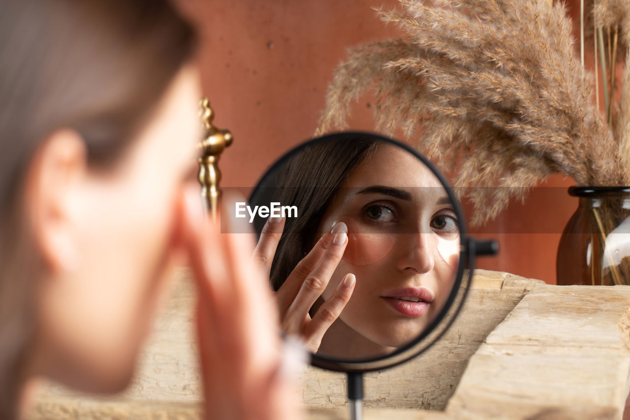 Mirror reflection of woman applying under-eye patch at dressing table closeup