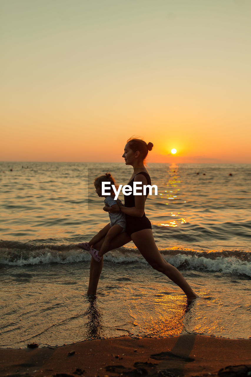 Mother with daughter wading in sea during sunset