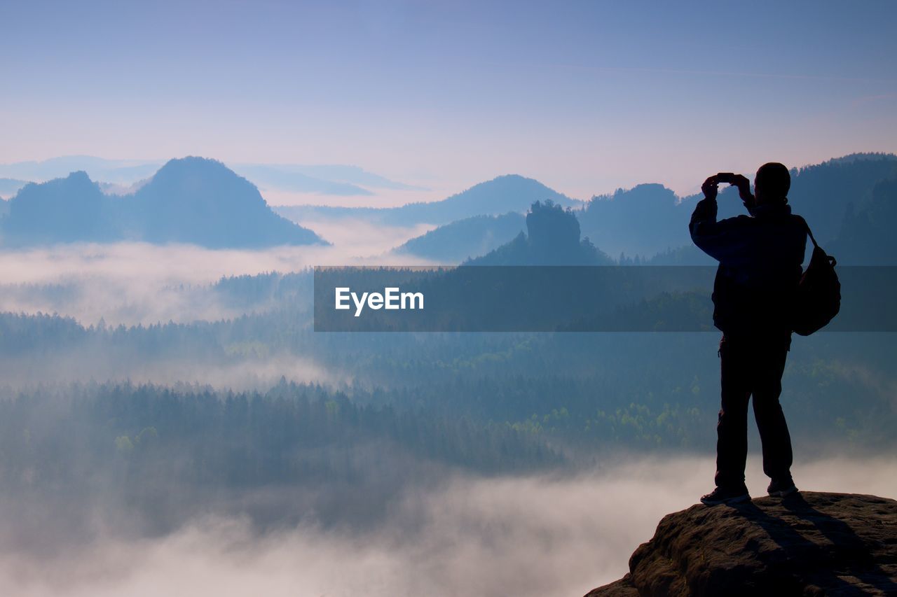 Tourist with backpack takes photos with smart phone on the rocky peak. dreamy fogy valley below