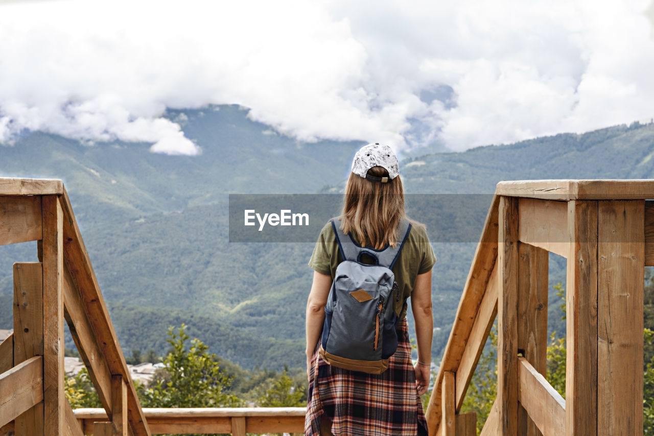 Rear view of woman in cap with backpacklooking at view of mountains and cloudy sky on viewpoint