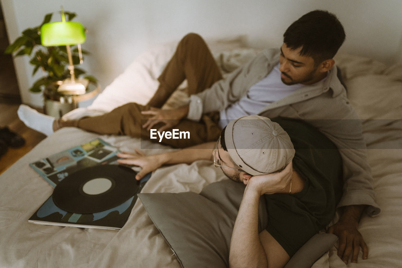 Gay couple with vinyl record talking to each other while lying on bed at home