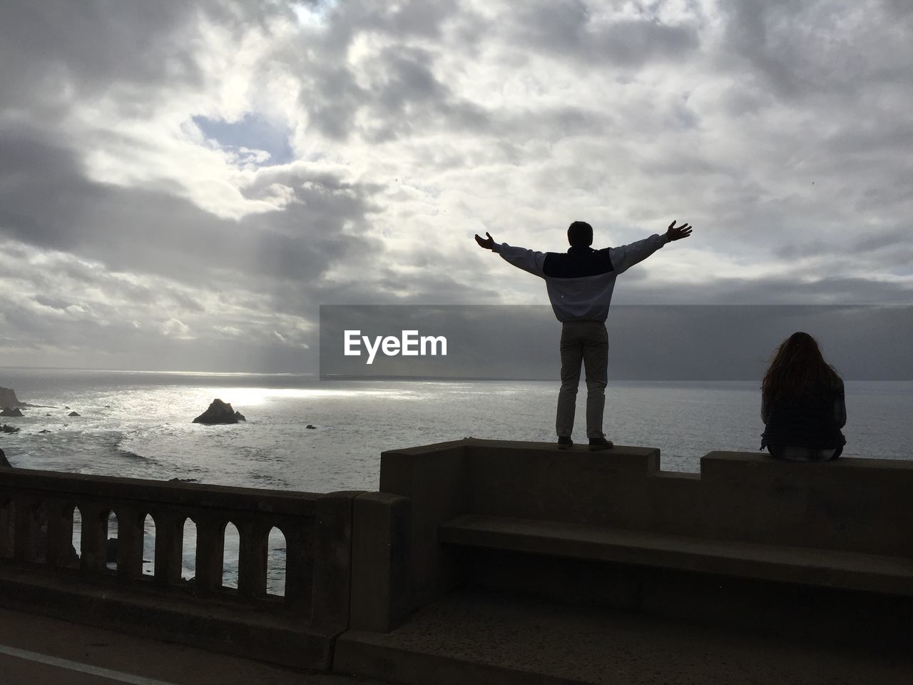 Man standing while woman sitting on retaining wall by sea against cloudy sky