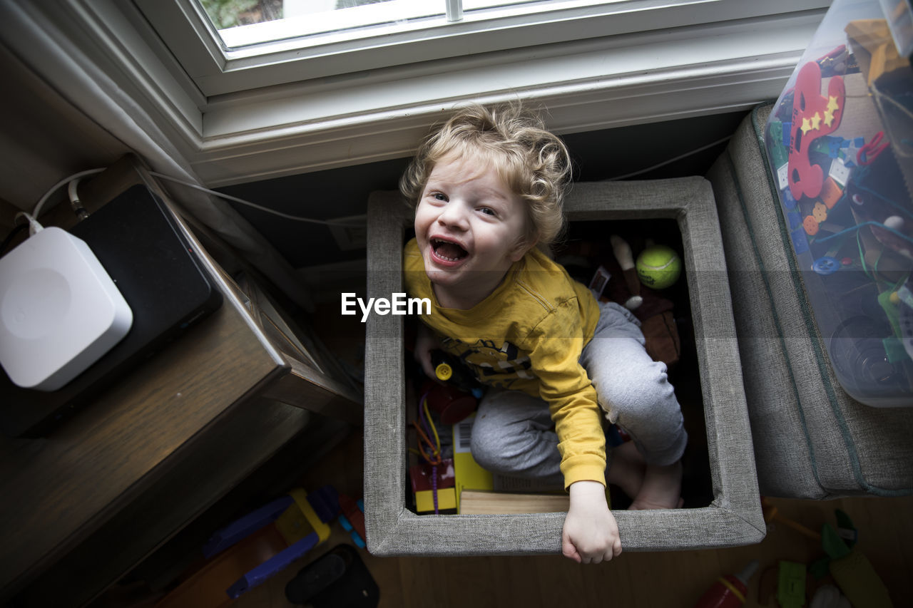 Laughing boy sits in toy box next to storage bins looks up at camera