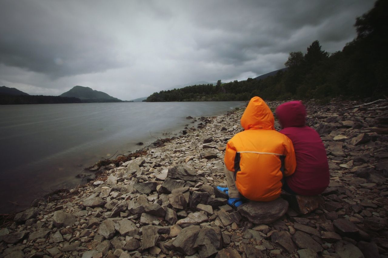 Rear view of man and woman in jacket sitting on rocks by lake against sky