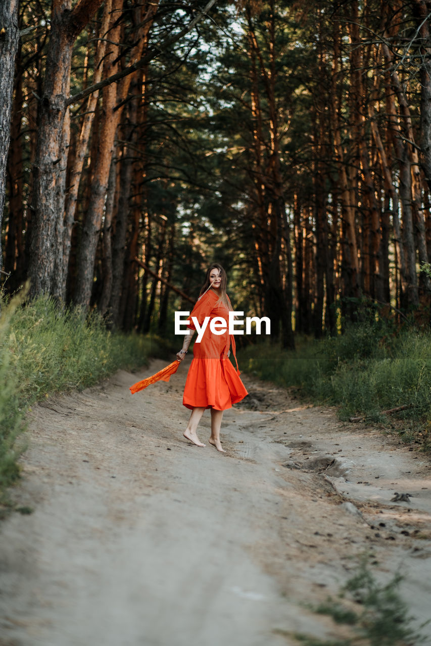 Barefoot happy young woman in red dress with hand raised dancing in pine forest at summer day.