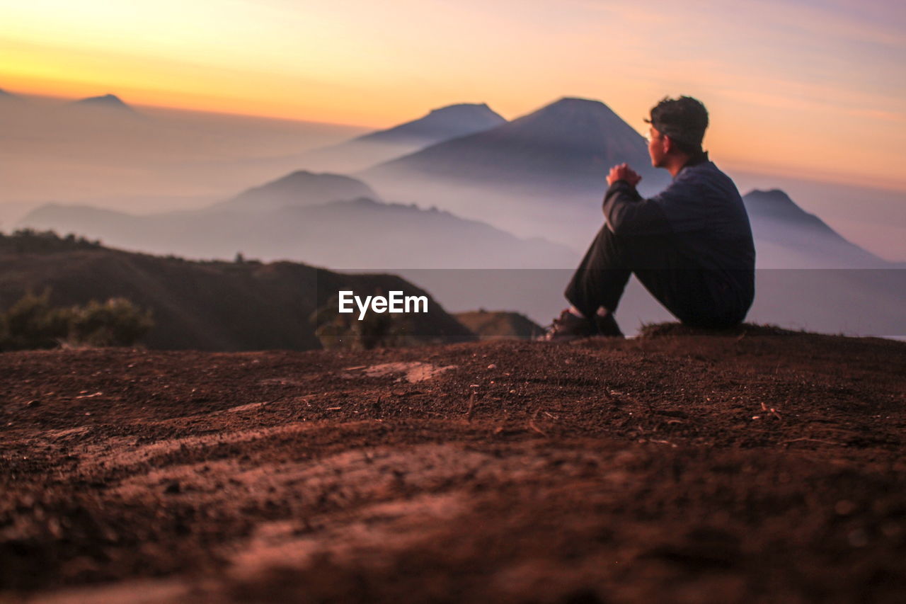 Man sitting on mountain against sky during sunset