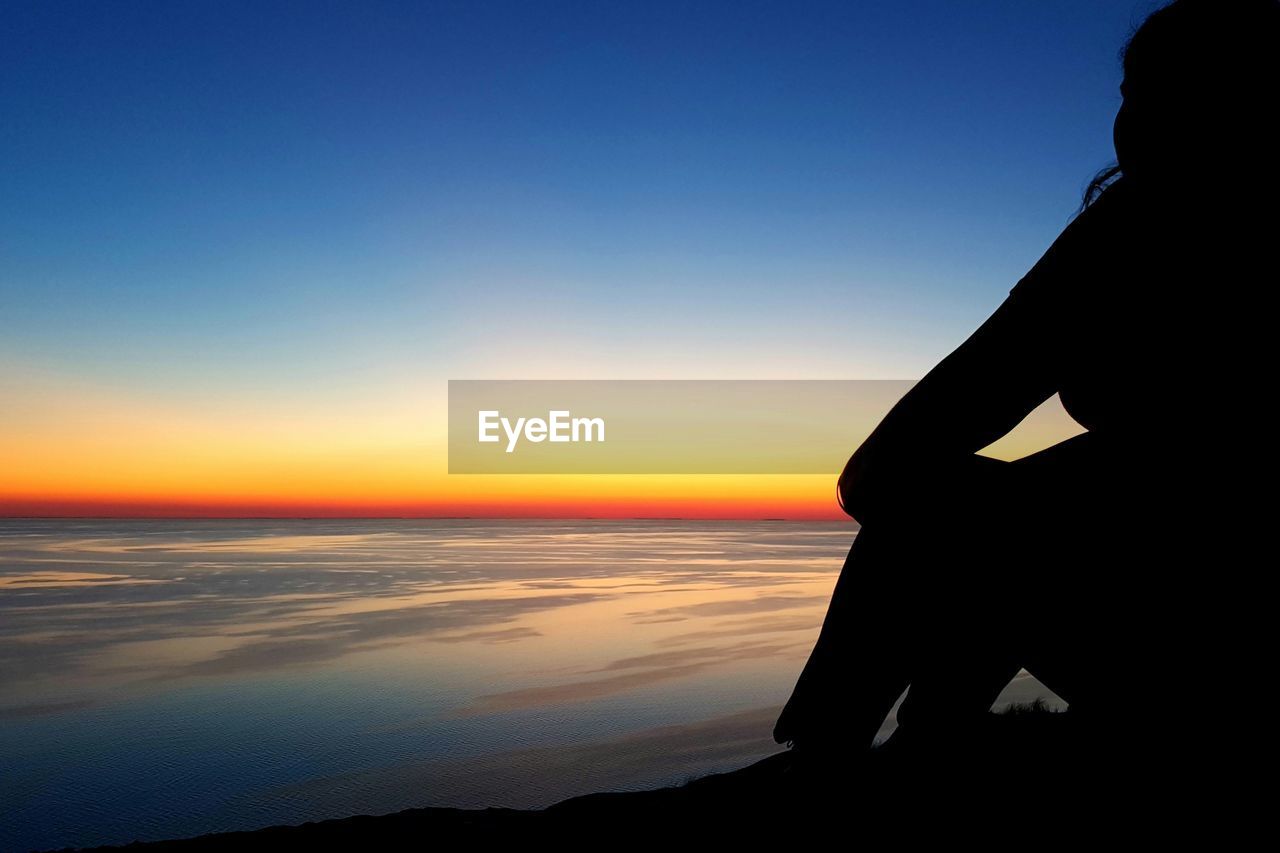 Silhouette woman sitting at seashore against clear sky during sunset