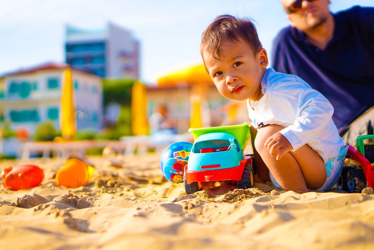 Portrait of baby boy playing with toys at beach while father in background