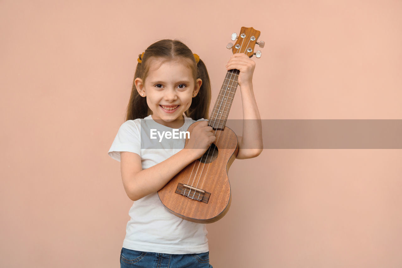 child, childhood, musical instrument, portrait, smiling, music, looking at camera, happiness, studio shot, string instrument, indoors, colored background, one person, guitar, emotion, cheerful, copy space, musician, holding, arts culture and entertainment, female, women, front view, musical equipment, enjoyment, casual clothing, plucked string instruments, cute, standing, person, positive emotion, fun, cut out, toddler, waist up, performance, lifestyles, three quarter length